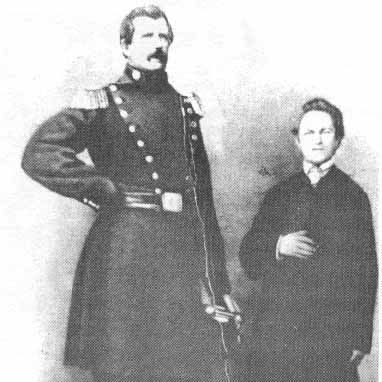 A black-and-white photograph of a tall man, Angus MacAskill, standing with his hand on his hip, wearing a military-style uniform with epaulettes and a belt, holding a sword. Beside him stands a shorter man in a dark suit with his hand on his chest. The background is plain and dark.