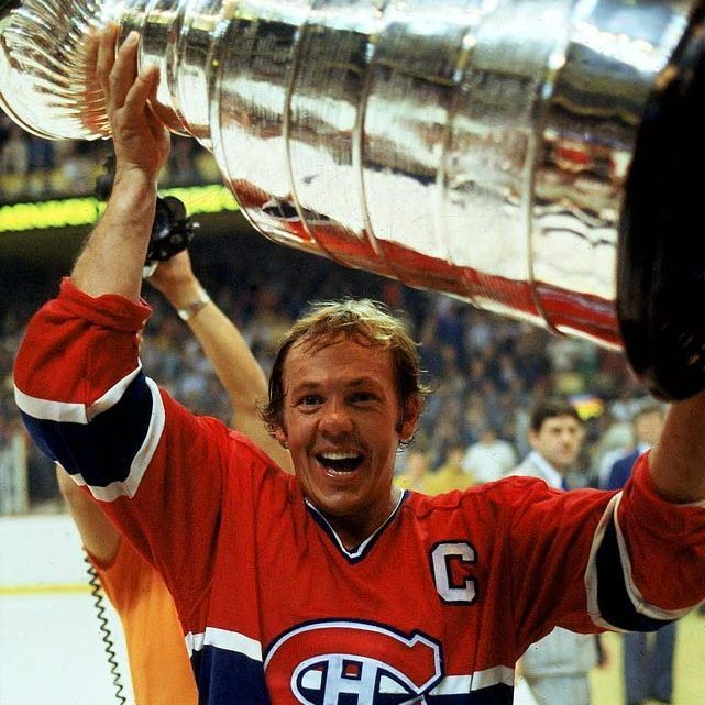 A hockey player wearing a red jersey with the "CHC" logo and a captain's "C" hoists the Stanley Cup trophy above his head, celebrating on the ice with a crowd and officials in the background.