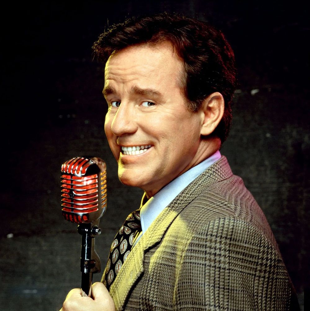 A person in a plaid suit jacket and patterned tie holding a vintage red and silver microphone, standing against a dark background.