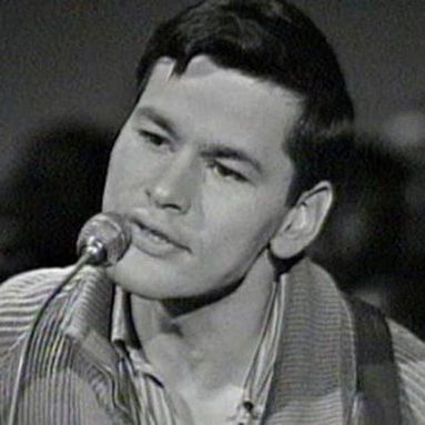 Black and white close-up photograph of Willie Dunn, a man with short dark hair combed to the side, looking slightly downward with his mouth open as if singing or speaking into a small microphone positioned in front of him. He is wearing a ribbed sweater over a collared shirt, with a guitar strap visible over his shoulder. The background is blurred, featuring indistinct figures that appear to be an audience.