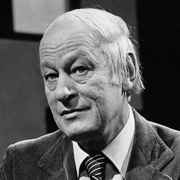 Black and white photo of René Lévesque, an older man with white hair, wearing a suit and striped tie, sitting and looking slightly to the side against a dark background with a checkered pattern.