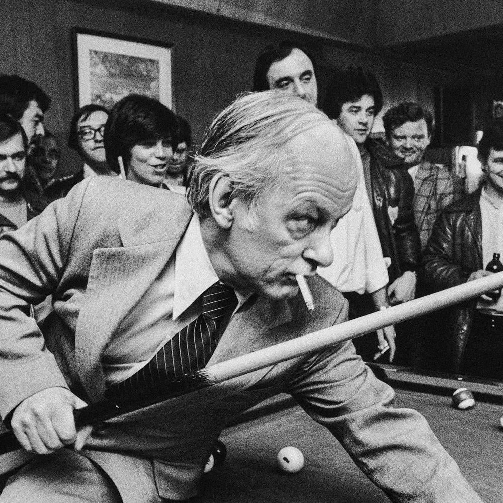 Black and white photo of Rene Levesque, wearing a suit and tie, playing pool with a cigarette in his mouth, leaning over a table with billiard balls. A group of people, some in leather jackets, watches in the background inside a room with wood-paneled walls and a framed picture.