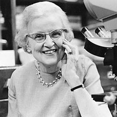 A black-and-white photograph of an elderly woman with glasses, smiling and holding a telephone receiver to her ear. She is wearing a necklace and a bracelet, and is dressed in a light-colored outfit. In the background, there is a microscope and other scientific equipment, suggesting a laboratory setting.