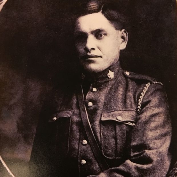 A black-and-white portrait of a man in a military uniform with a maple leaf insignia on the collar, featuring a buttoned jacket with pockets and a leather strap across the chest. He holds a sword or similar object in his right hand and gazes slightly to the side, with a decorative frame surrounding the image.