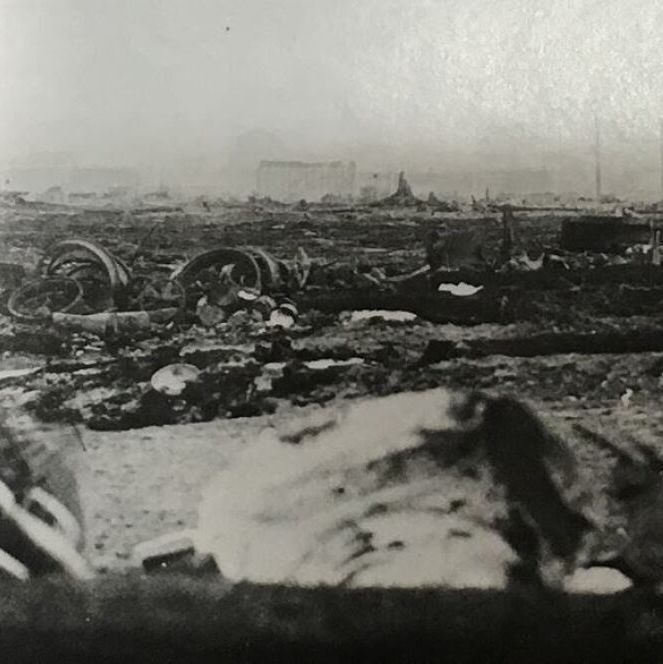 The black-and-white photograph depicts the aftermath of the Matheson Fire of 1916 in northern Ontario, Canada. It shows a desolate, ash-covered landscape stretching into the distance, with scattered debris including charred wheels, axles, and metal remnants from destroyed vehicles or rail cars in the foreground. In the background, faint outlines of ruined buildings and structures stand amid the widespread devastation, under a hazy sky.