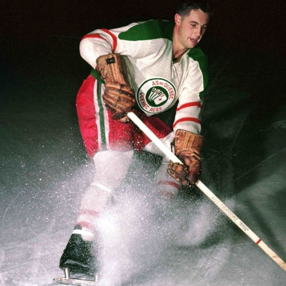 The photo shows Jean Béliveau in action on the ice, wearing a Quebec Aces hockey jersey with a circular logo featuring the text 