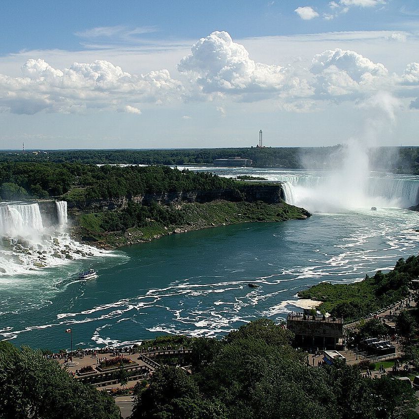 A scenic view of Niagara Falls, featuring three separate waterfalls surrounded by lush green forests and a clear blue sky with scattered clouds. The river below flows turbulently, with a boat navigating the waters and people visible on observation decks and pathways.