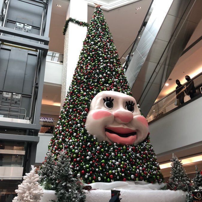 A large Christmas tree in a shopping mall atrium, decorated with red, green, silver, and white ornaments. The tree features a giant animated face with big eyes, rosy cheeks, and a wide smiling mouth; this is Woody the Talking Tree. Smaller snow-dusted trees surround the base. People stand on upper balconies overlooking the display.