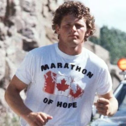 The photo shows a man with curly hair wearing a white T-shirt with "Marathon of Hope" and a design featuring multiple Canadian maple leaf flags printed on it. He appears to be running or jogging outdoors, with a focused expression. In the background, there is a police car with its lights on, parked near a rocky landscape and some greenery. The scene suggests a determined effort, likely related to Terry Fox's famous cross-Canada run to raise awareness for cancer research.