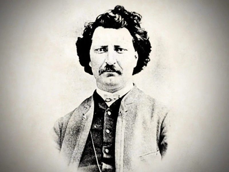 The image depicts a black and white photograph of a man from the 19th century. He has dark, wavy hair and a mustache, and is wearing a high-collared shirt with a buttoned jacket over it. The photograph has a vintage, historical appearance, suggesting it might be from the mid to late 1800s.