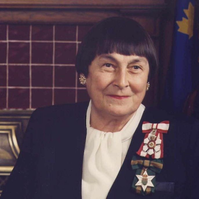 A photograph of a person wearing a dark suit jacket and a white blouse, adorned with multiple medals and ribbons on the left side, including a red and white ribbon with a star and a green and white ribbon with a star. The individual is also wearing earrings and is seated in front of a wooden panel with a tiled maroon background and a flag featuring a blue field with yellow and red symbols.