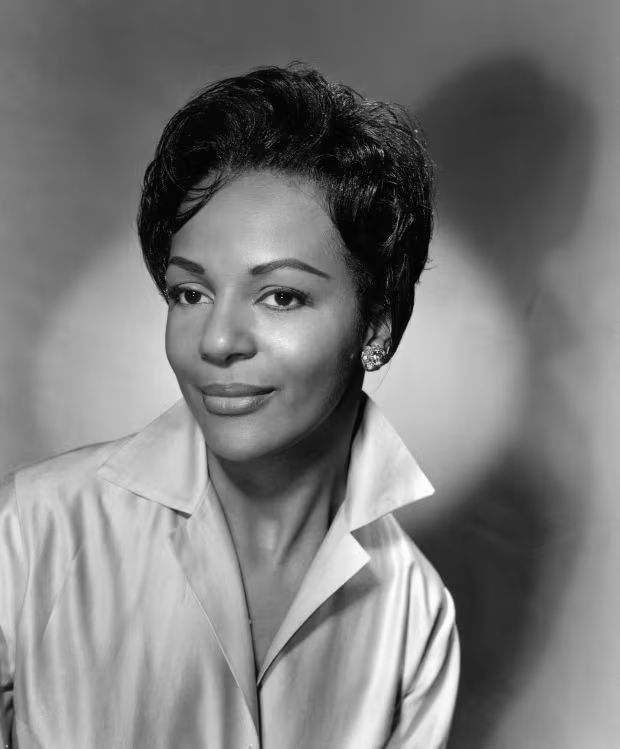 The photo is a black-and-white portrait of a woman with short, wavy hair styled neatly. She is wearing a collared shirt with a pointed collar, giving a classic and elegant appearance. She has large, expressive eyes and is adorned with noticeable earrings, possibly stud or pearl, adding a touch of sophistication. The background is softly blurred, creating a focus on her face, and the lighting highlights her facial features, giving the image a timeless, vintage feel. Her expression is calm and composed, with a subtle smile, exuding confidence and poise. The overall tone of the photo suggests it could be from a mid-20th-century portrait session, possibly for a professional or promotional purpose.