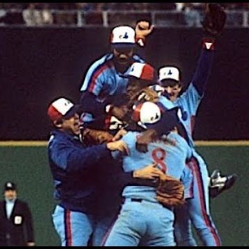 A group of Montreal Expos players in light blue uniforms with red and white accents celebrate on the field, piling into a joyful huddle with arms raised in triumph. One player wears jersey number 8 on the back. An umpire in black stands nearby in the background.