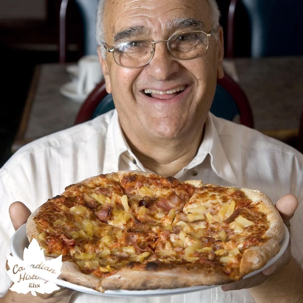 
This photo shows a person holding a large pizza with a variety of toppings, including what appears to be ham and pineapple, suggesting it might be a Hawaiian pizza. The individual is smiling broadly, indicating enjoyment or pride in presenting the pizza. The setting appears to be indoors, possibly in a restaurant or a dining area, as there are chairs and a table visible in the background. The pizza is held on a white plate, and the person is wearing a light-colored shirt.