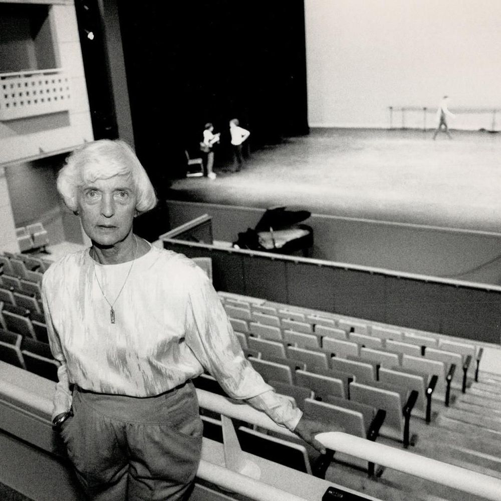Black and white photograph of an older woman with short white hair, wearing a light silky blouse, pants, a pendant necklace, and a wristwatch, standing in an empty auditorium and leaning on a railing with a serious expression as she gazes toward the stage. In the background, a vast empty stage with a black curtain features a grand piano, a ballet barre along the wall, and three distant figures: two people conversing near a chair (one possibly holding an instrument or object), and another person walking across the floor. Rows of unoccupied seats fill the midground.