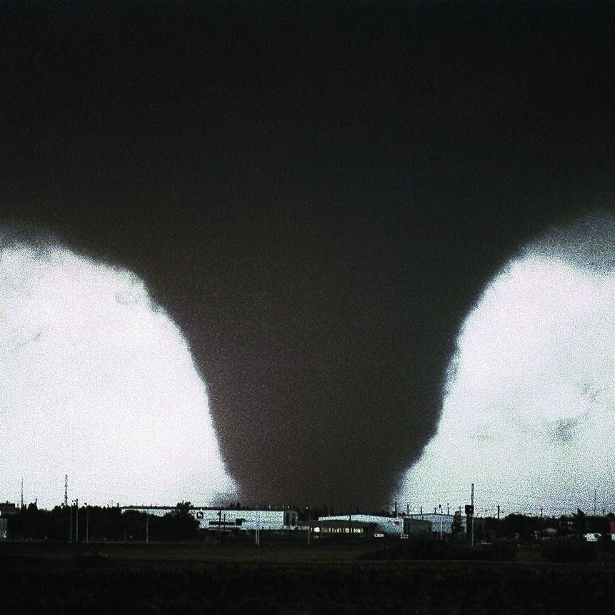 The provided photo shows the iconic image of the 1987 Edmonton tornado, captured by photographer Steve Simon. It depicts a large, wedge-shaped funnel cloud descending from a dark storm sky, touching down over an industrial area known as Refinery Row in eastern Edmonton, Alberta. The tornado appears as a bright, contrasting formation against the blackened background, with buildings, utility poles, and structures visible in the foreground.