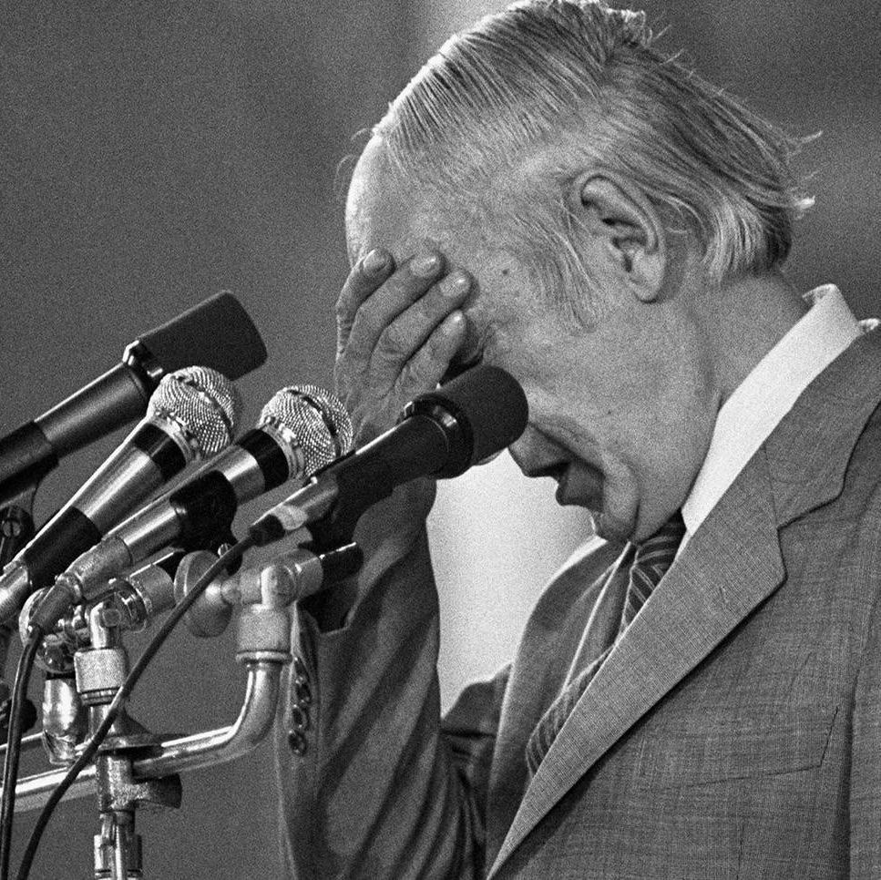 Black and white photo of René Lévesque, a man with white hair, wearing a suit and tie, standing at a podium with multiple microphones, holding his head with one hand in a gesture of exhaustion or frustration.