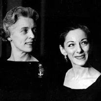 Black and white photograph of two women in formal off-the-shoulder dresses, one wearing a brooch and both with earrings, looking to the side with subtle smiles.