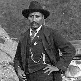 A black-and-white photograph of a man wearing a dark suit, vest, and tie, with a pocket watch chain visible. He has a hat tilted to one side and stands with one hand on his hip and the other holding a cigarette, with a rocky background.