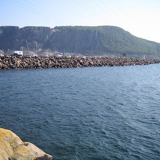 A color photo of a long, straight rocky causeway extending across calm blue water toward a steep, tree-covered cliff in the background. The top of the causeway is lined with a long row of parked cars, trucks, and a camper van, with a few people standing nearby. Power lines stretch overhead, and the scene is bright under a clear sky. The foreground shows water lapping against large shoreline rocks.