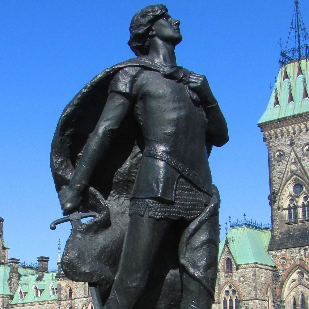 A bronze statue of a historical figure in medieval attire, including a tunic, belt, and cape draped over one shoulder, stands outdoors. The figure holds a sword in one hand and gazes upward. In the background, a large Gothic-style building with green copper roofs and pointed towers is visible under a clear blue sky.