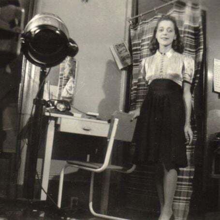 The black-and-white photo depicts a vintage hair salon or beauty parlor setting. A person stands in the center, wearing a short-sleeved blouse with a pleated front and a dark skirt. To the left, there is a hairdryer with a large hood attached to a stand, positioned next to a small desk with a drawer and various items on it. A chair is placed in front of the desk. The background features striped curtains, a wall-mounted light bulb, and a small table or chair with additional equipment. The overall ambiance suggests a mid-20th-century interior.