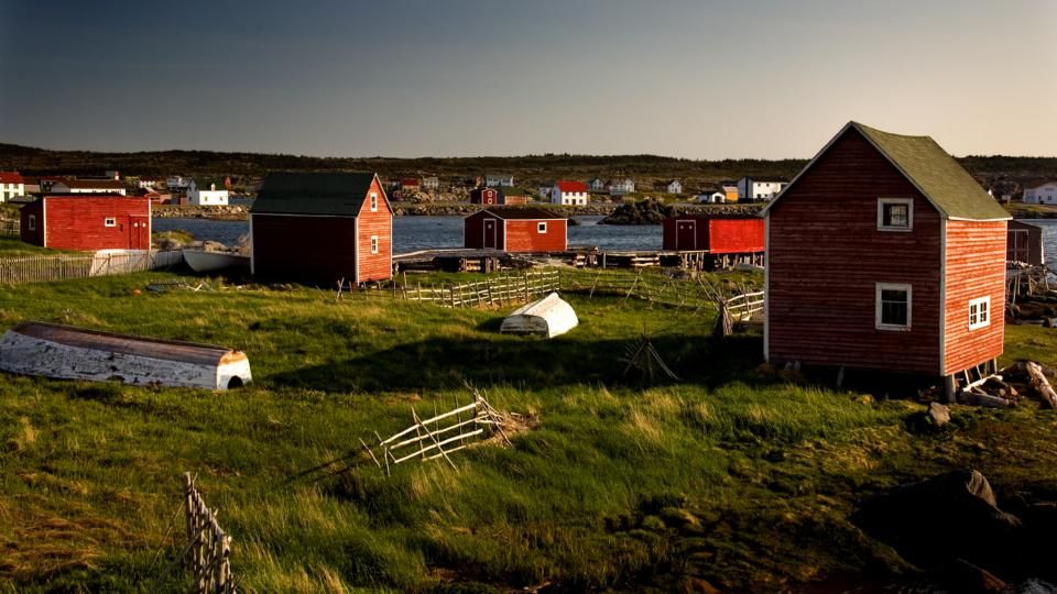 The color photo depicts a coastal village with several red wooden houses and sheds on a grassy landscape. Small boats are scattered around, and the scene is set near a body of water with more buildings in the background under a clear sky.