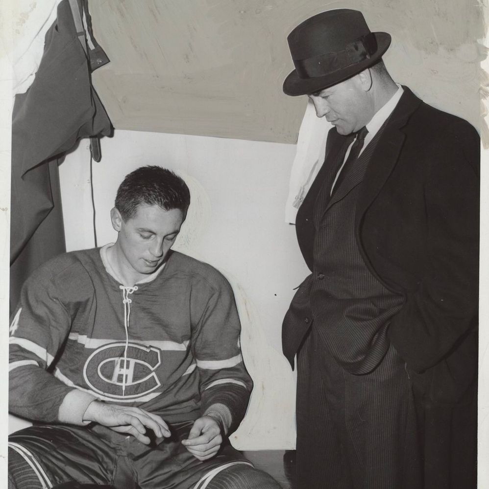 The photo shows Jean Béliveau sitting on a bench in a Montreal Canadiens hockey jersey with the number 4 and the team's logo on the chest. He is looking down at his hands, wearing full hockey gear including pants and skates. Standing next to him is a man in a suit and hat, possibly a coach or official, looking down at him, with a coat hanging on the wall in the background.