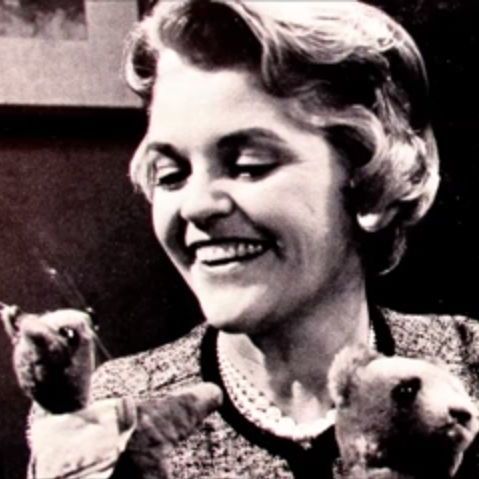 A person with short, wavy hair wearing a suit and pearl necklace smiles while holding two hand puppets, one resembling a mouse, against a dark background.