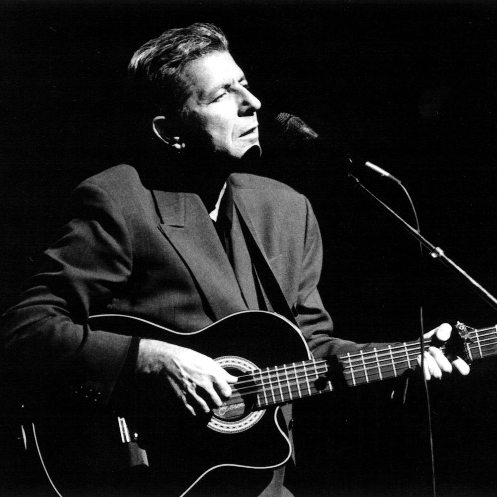 A black-and-white photograph of Leonard Cohen, a man with short hair, playing an acoustic guitar and singing into a microphone on stage, dressed in a dark suit jacket.