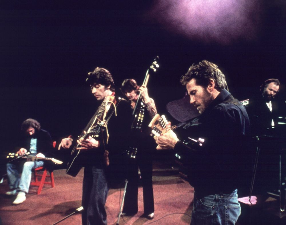 This image captures a live performance by a band, likely from the 1970s or 1980s, given the style of clothing and instruments. The band members are playing electric guitars, with one musician using a double-neck guitar, which was popular among rock guitarists of that era. The setting appears to be a stage with dim lighting and a spotlight effect, typical of rock concerts during this period. The audience is not visible, focusing the viewer's attention entirely on the performers and their interaction with their instruments.