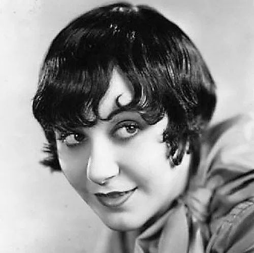 A black-and-white portrait of a woman with short, wavy bobbed hair typical of the 1920s, wearing a high-collared blouse with a loosely tied bow at the neck. She gazes slightly to the side with a subtle, knowing smile, exuding classic silent film-era elegance against a plain backdrop.