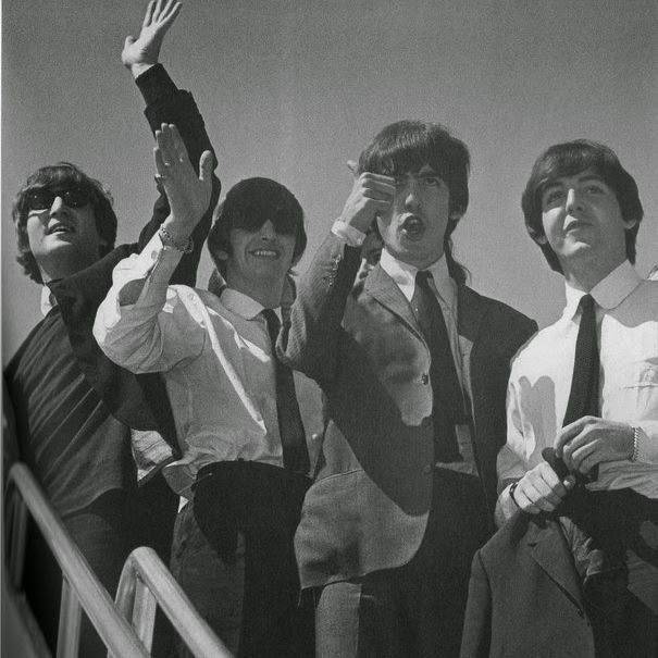 Four members of the Beatles—John Lennon, Paul McCartney, George Harrison, and Ringo Starr—standing on an airplane staircase, dressed in suits and ties from the 1960s. John Lennon, on the left, wears sunglasses and raises his left hand in a wave, while Paul McCartney, second from the left, smiles and waves with his right hand. George Harrison, second from the right, makes a playful face with his mouth open and hand near his face, and Ringo Starr, on the right, holds his jacket and looks forward. The black-and-white photo captures them against a clear sky, with a metal railing visible in the foreground.