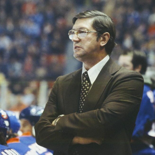 A vintage photograph of a man in a suit and tie, standing with arms crossed, on the sidelines of an ice hockey game. He is wearing glasses and a serious expression, with a blurred crowd and hockey players in blue and orange uniforms in the background.