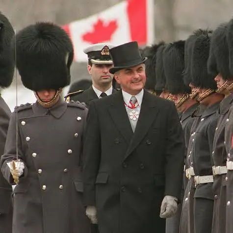 A man wearing a black top hat, black overcoat, white gloves, and a red-and-white neck medal walks past a line of soldiers in dark winter dress uniforms with tall black bearskin hats, inspecting the troops. A Canadian flag is visible in the background. The scene appears to be a formal military ceremony on a cold day.
