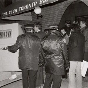This photo appears to be a black-and-white image taken outside a club in Toronto, as indicated by the sign "THE CLUB TORONTO 231" above the entrance. The scene shows a group of people gathered near the entrance of the club, with some individuals wearing jackets that suggest it might be cold outside. There is a police vehicle, specifically an armored or riot control vehicle, parked in front of the club. The vehicle has the word "POLICE" visible on it, and its license plate reads "AB5-538."