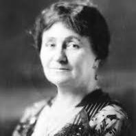 A black-and-white photograph of a woman with short, dark hair, wearing a patterned dress with a high neckline and lace details. She is posed against a plain background.