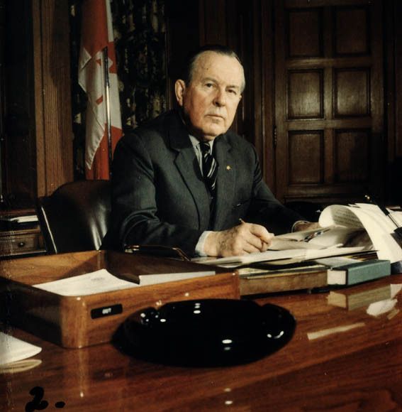A black-and-white photo of a man in a suit sitting at a wooden desk in an office, holding a pen and reviewing documents. The desk has various items, including an ashtray, papers, and books. A Canadian flag is visible in the background, and the room features wood-paneled walls.