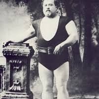 A black-and-white photograph of a muscular man standing with one hand resting on an ornate pedestal. He is wearing a sleeveless athletic outfit, a belt, and sandals, with a painted backdrop of trees behind him.