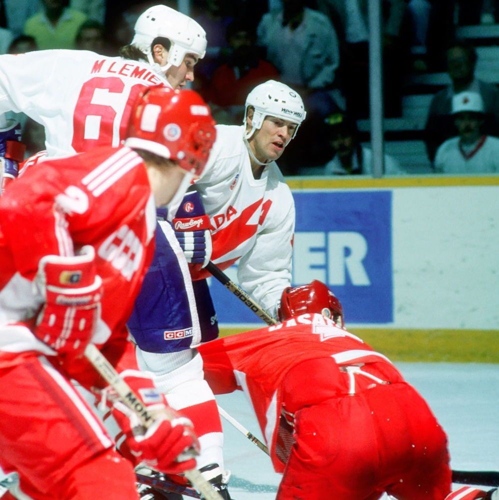 A color photograph of an intense hockey game moment on the ice, featuring players in action. A player in a white jersey with 