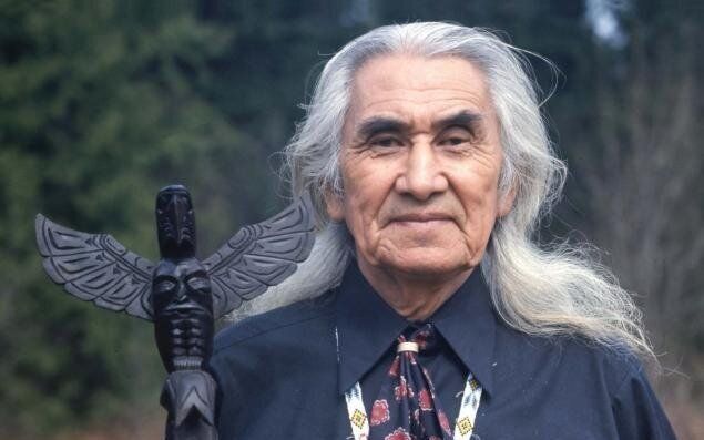 The photo shows an individual with long, white hair, wearing a dark shirt and a patterned tie with a beaded medallion necklace. They are holding a carved wooden staff topped with a totem-like figure, featuring a face and outstretched wings, possibly representing an eagle or another significant symbol. The background is an outdoor setting with blurred greenery, suggesting a forest or wooded area. The overall composition suggests a cultural or ceremonial context, possibly related to Indigenous traditions.