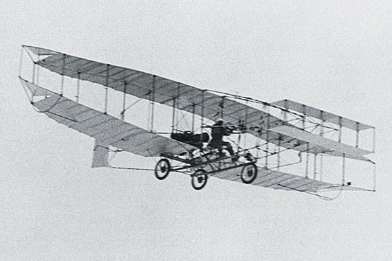 The black-and-white photo shows an early biplane in flight, with two sets of wings stacked vertically and a person piloting it. The aircraft has a lightweight frame, visible wheels, and a simple design typical of early 20th-century aviation, set against a plain sky background.