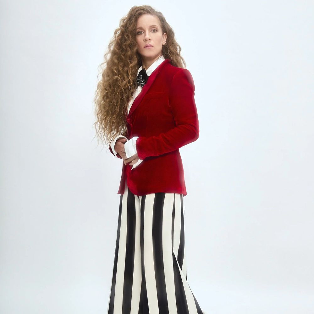 A person standing against a plain white background, wearing a red velvet blazer over a white shirt with a black tie, and wide-leg pants with bold black and white vertical stripes. The individual has long, curly hair and is posed with hands clasped in front.