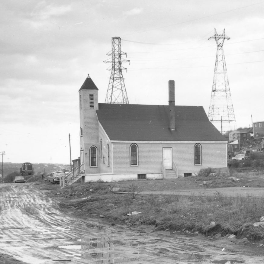 A black-and-white photograph depicting a small town with a prominent church featuring a steeple and arched windows in the foreground. The church is situated along a muddy, unpaved road with visible tire tracks and puddles. In the background, there are power lines supported by tall utility poles, and a hillside with numerous modest houses. A few cars are parked along the road, and a person is sitting on a bench near the right side of the image. The sky is overcast with scattered clouds.