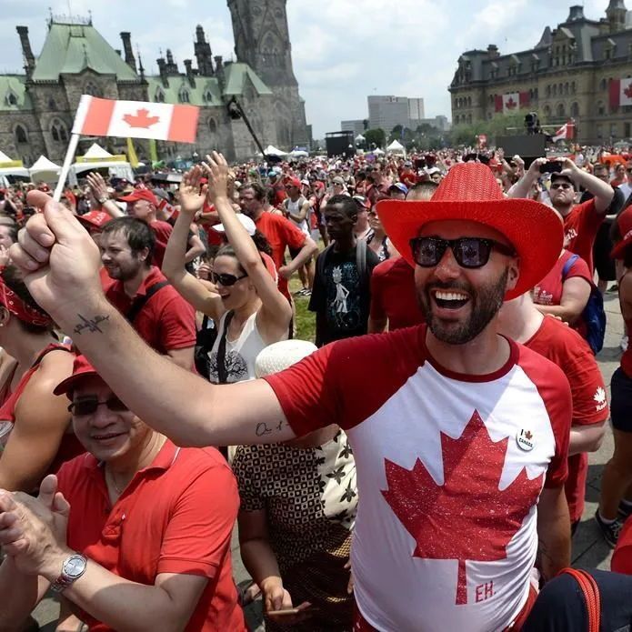 The photo depicts a lively outdoor celebration with a large crowd of people, many wearing red and white clothing and accessories featuring the Canadian flag. The central figure is a man wearing a red cowboy hat and sunglasses, smiling broadly, and dressed in a shirt with a large maple leaf design and the word "EH" on it. He is holding a small Canadian flag raised in his right hand. The background shows a grand building with green roofs and Canadian flags, suggesting a significant national event, likely Canada Day, given the date and context. Other people around him are also waving flags and cheering, creating a festive atmosphere.