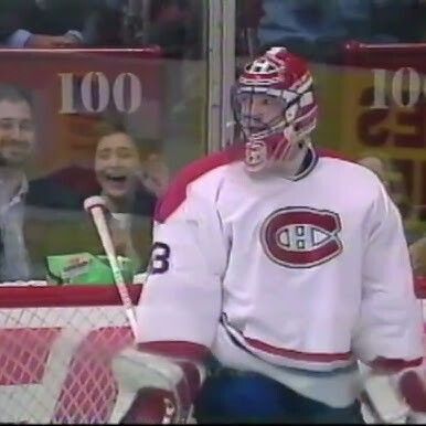 A hockey goalie in a white Montreal Canadiens jersey with the red and blue CH logo, wearing a red mask, stands in front of the net looking toward the camera with a serious expression. Behind the glass directly in front of him, a young fan in the crowd is screaming excitedly with mouth wide open and hands pressed against the glass. A sign with "100" is visible in the background.