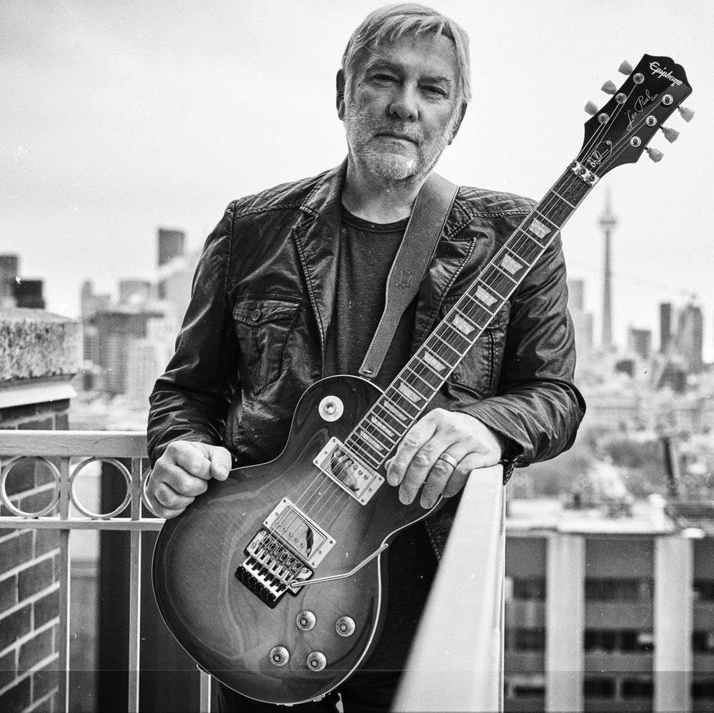 Black and white photo of a man with a beard and light hair, wearing a leather jacket, holding an electric guitar on a balcony with a city skyline, including tall buildings and a distinctive tower, in the background. The image has a dated timestamp from March 25, 1999.