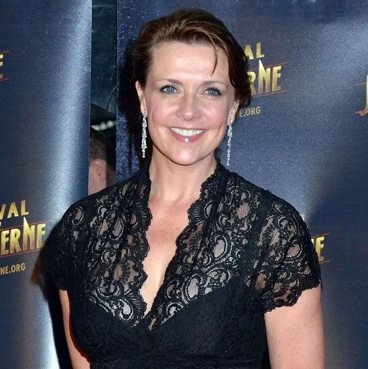 A woman with short brown hair wearing a black lace dress with a deep V-neckline and long, dangling earrings, smiling at a red carpet event. The backdrop features the text 