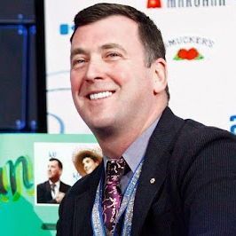 A man with short dark hair, wearing a dark suit jacket, light blue dress shirt, and patterned tie, smiles broadly at the camera while seated at a table. A large green nameplate in front of him reads "Brian" in stylized yellow letters, with smaller photos on it. Behind him is a white backdrop with logos including "Muckers" and a red rose emblem.