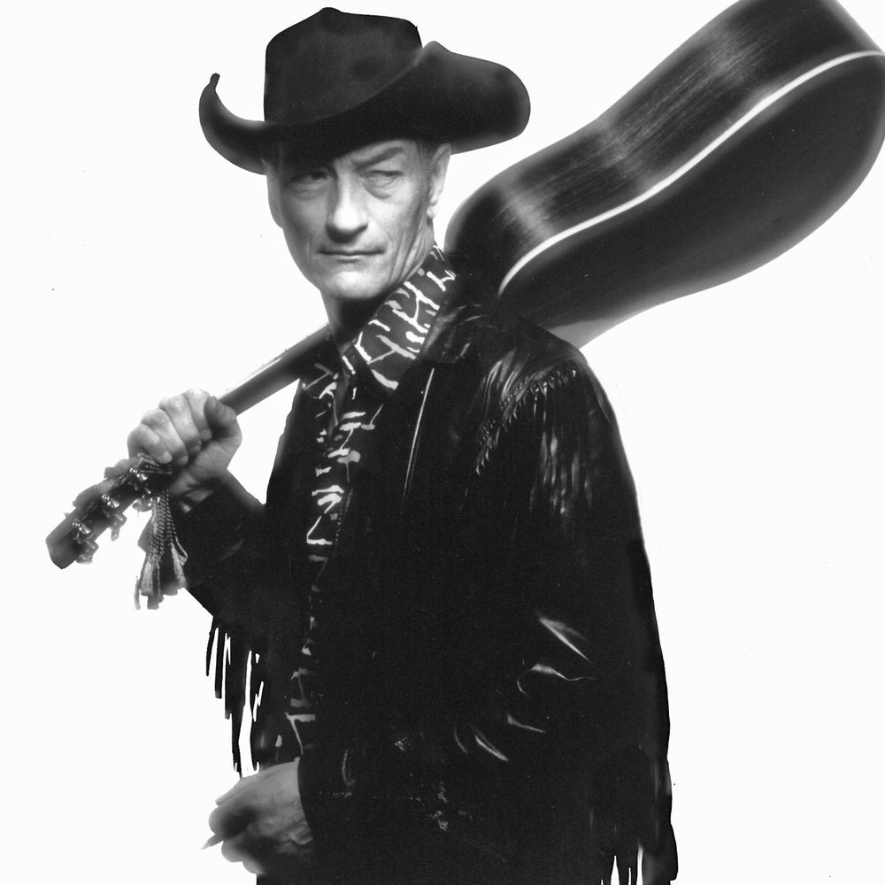 The black-and-white photo features a person wearing a wide-brimmed cowboy hat and a fringed leather jacket. They are holding a guitar over their shoulder, with one hand gripping the neck and the other resting near the body of the instrument. The individual has a patterned shirt underneath the jacket. The background is plain white, keeping the focus on the subject.